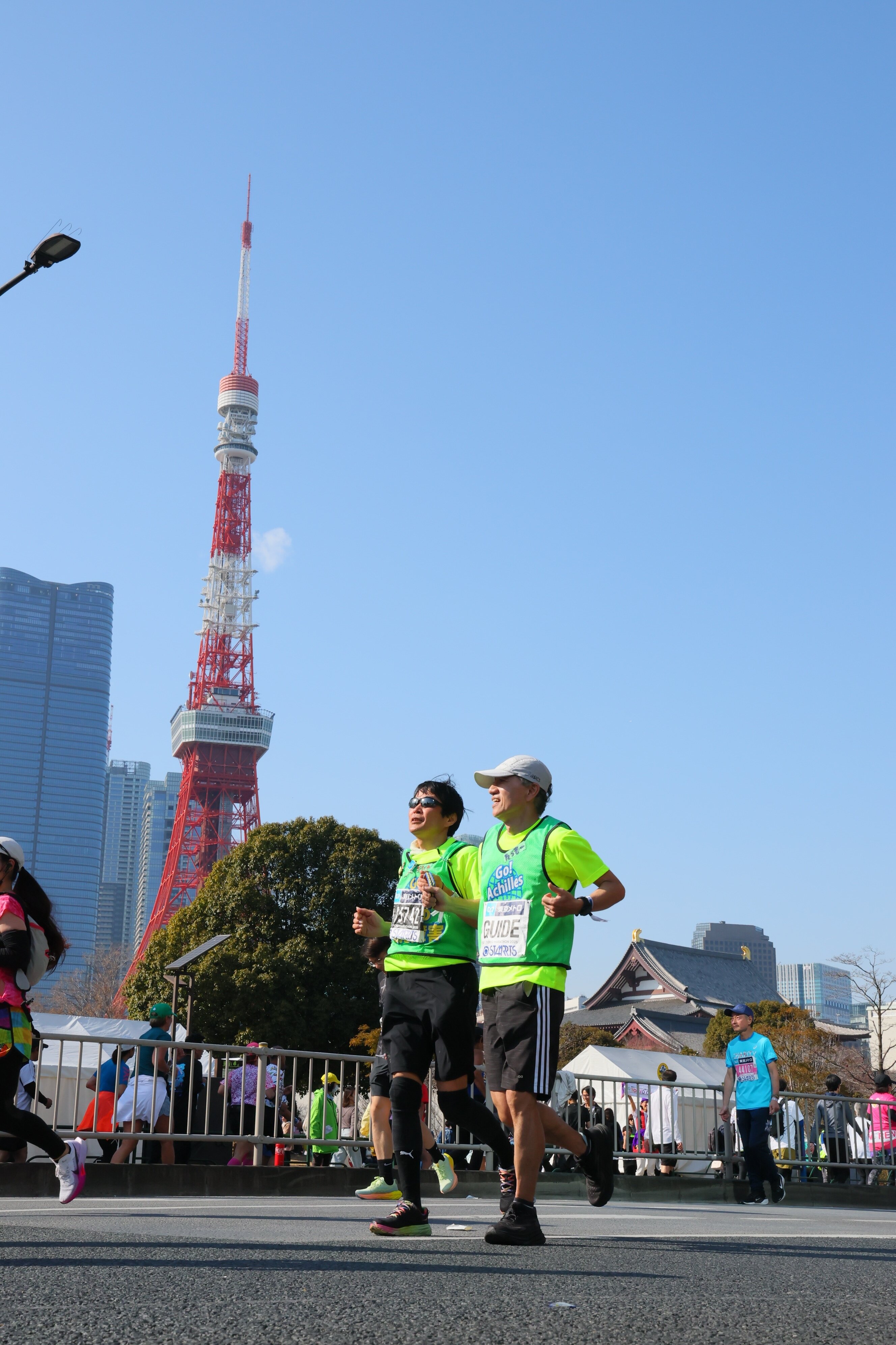 画像には、マラソン大会の様子が写っています。 手前には、ランナーたちが道路を走っている様子が捉えられています。男性2人が並んで走っており、蛍光色のグリ.jpg